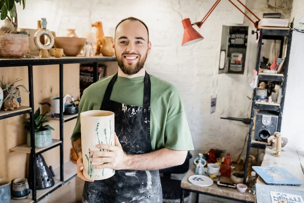 Smiling bearded artisan in apron holding clay vase and looking at camera in ceramic workshop — Stock Photo