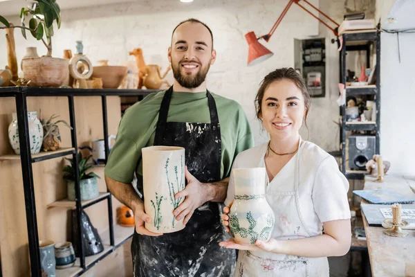 Улыбающаяся пара ремесленников в фартуках, держащих глиняные вазы и смотрящих в камеру в керамической мастерской — стоковое фото