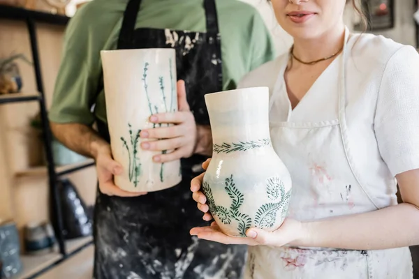 Vista cortada de artesã em avental segurando vaso de barro perto namorado borrado na oficina de cerâmica — Fotografia de Stock