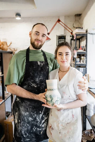 Sorrindo casal de artesãos em aventais abraçando e segurando vaso de argila em oficina de cerâmica no fundo — Fotografia de Stock