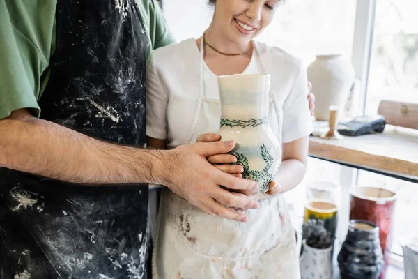 Vista recortada de la artesana sonriente sosteniendo jarrón de arcilla y de pie cerca de novio en el estudio de cerámica - foto de stock
