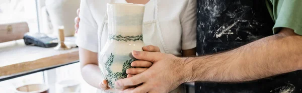 Vue recadrée d'un couple d'artisans dans des tabliers tenant un vase en argile dans un atelier de céramique, bannière — Photo de stock