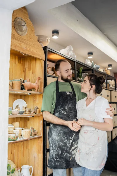 Couple of artisans in aprons holding hands near rack in blurred ceramic workshop at background — Stock Photo