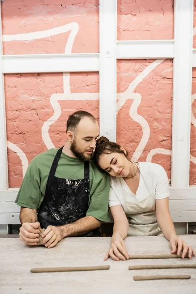 Craftsman in apron talking to smiling girlfriend and shaping clay on table in ceramic workshop — Stock Photo