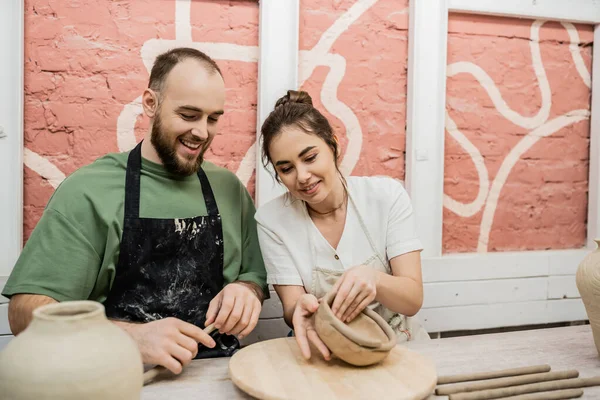 Artisanat souriant dans un bol en argile de mise en forme de tablier près du petit ami dans un atelier de céramique — Photo de stock