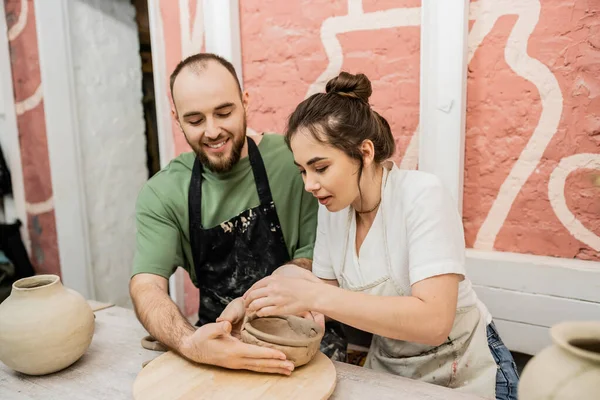 Artesano sonriente en delantal haciendo tazón de barro con novia en taller de cerámica - foto de stock
