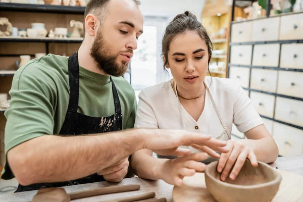 Bearded craftsman in apron shaping clay bowl and talking to girlfriend in ceramic workshop — Stock Photo