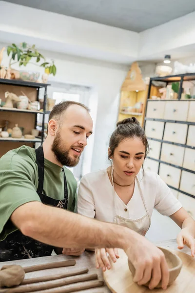 Smiling craftsman talking to girlfriend and taking clay bowl near girlfriend in ceramic studio — Stock Photo
