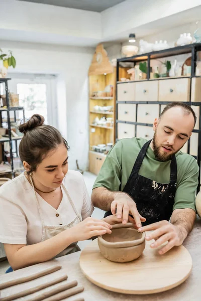 Couple of potters in aprons shaping clay bowl together while working in ceramic workshop — Stock Photo
