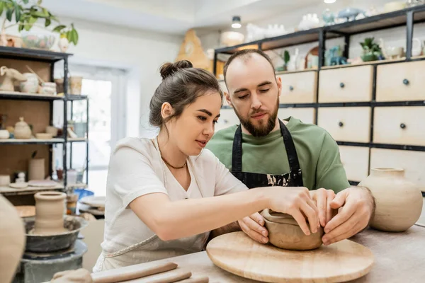 Брюнетка в фартуке делает глиняную миску с парнем вместе в керамической мастерской — стоковое фото