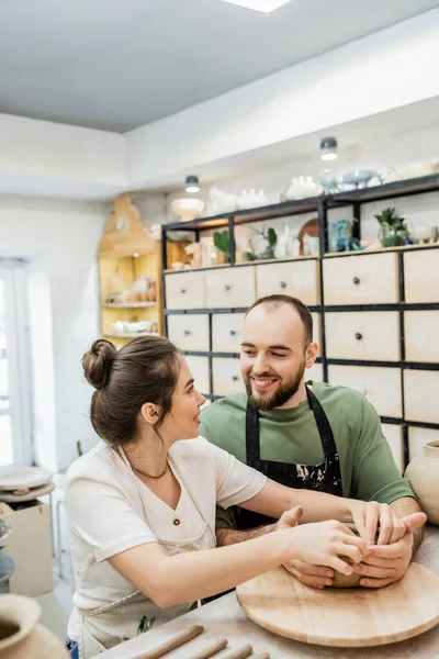 Sorrindo casal de artesãos em aventais fazendo tigela de barro e olhando uns para os outros no estúdio de cerâmica — Fotografia de Stock