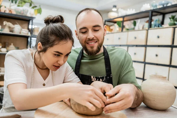 Smiling artisan in apron making clay earthenware with girlfriend while working in ceramic workshop — Stock Photo