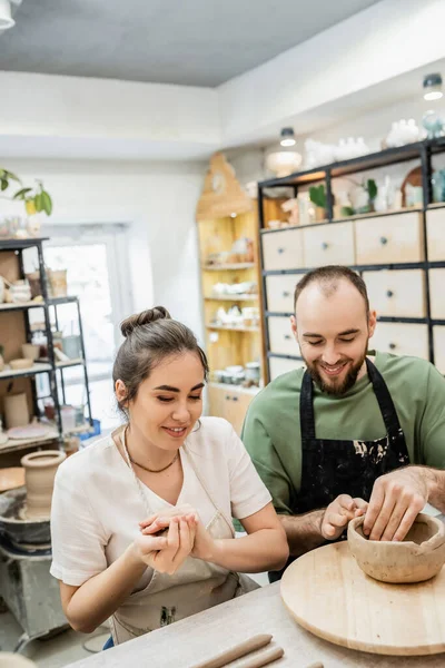 Smiling craftswoman in apron sitting near boyfriend shaping clay bowl on wooden board in workshop — Stock Photo