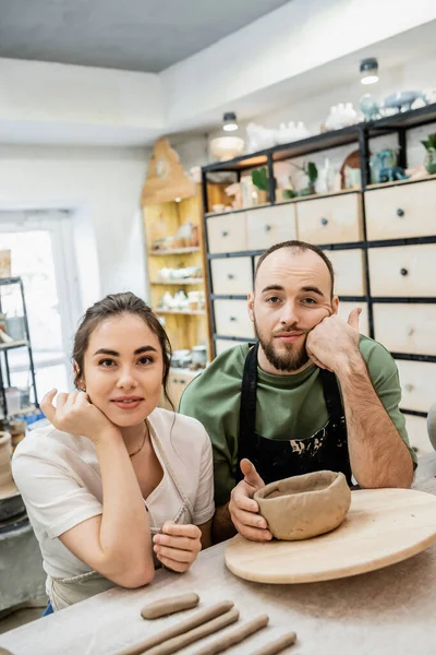 Artesã sorridente em avental olhando para a câmera perto de namorado triste e argila na oficina de cerâmica — Fotografia de Stock