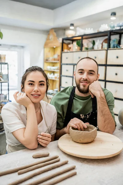 Cheerful couple of artisans in aprons looking at camera near clay on table in ceramic workshop — Stock Photo