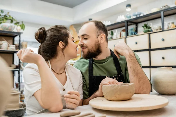 Smiling couple of potters in aprons kissing while sitting near clay in ceramic workshop — Stock Photo
