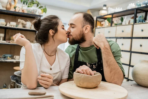 Romantic couple of artisans in aprons kissing near clay on table in ceramic studio at background — Stock Photo