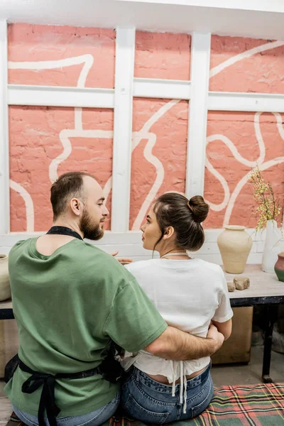Side view of couple of potters in aprons spending time in ceramic workshop at background — Stock Photo