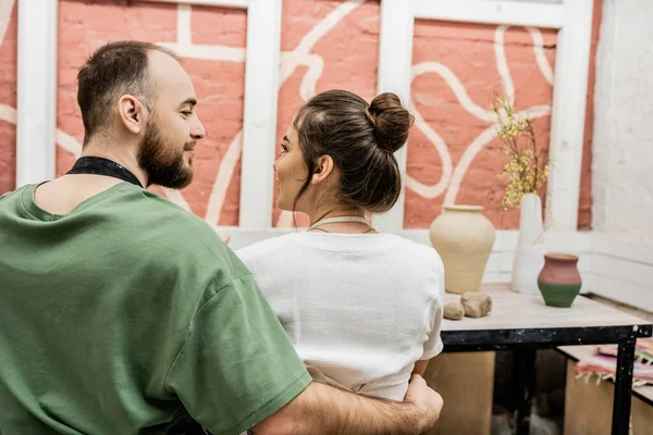 Bearded craftsman in apron hugging girlfriend while sitting near clay in ceramic workshop — Stock Photo