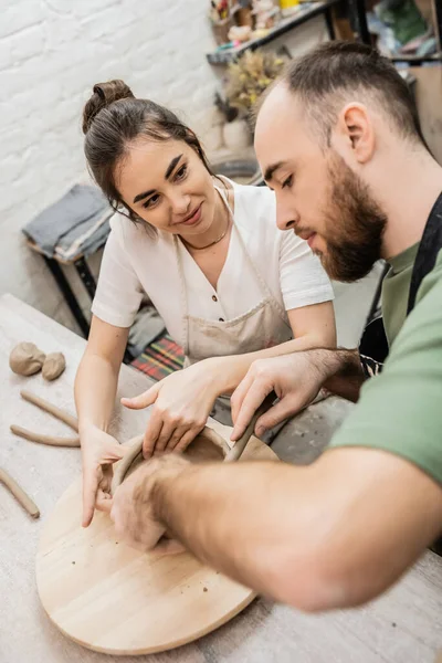 Positive craftswoman in apron shaping clay with blurred boyfriend together in workshop — Stock Photo