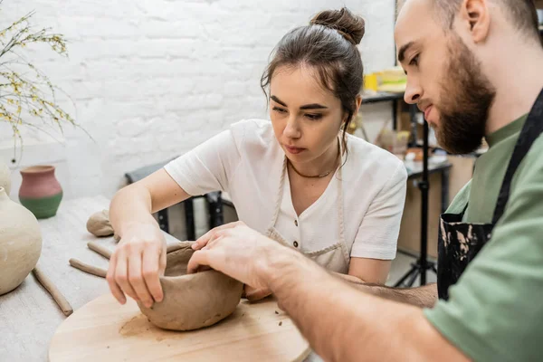 Brünette Kunsthandwerkerin in Schürze formt Tonschale mit bärtigem Freund in Keramik-Werkstatt — Stockfoto