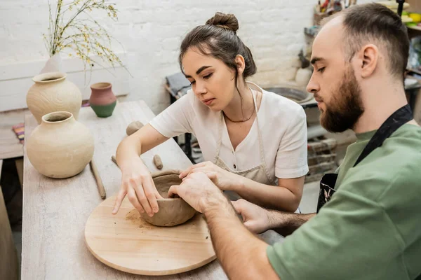 Casal de oleiros em aventais moldando tigela de argila enquanto trabalhava perto de vasos na mesa em oficina de cerâmica — Fotografia de Stock