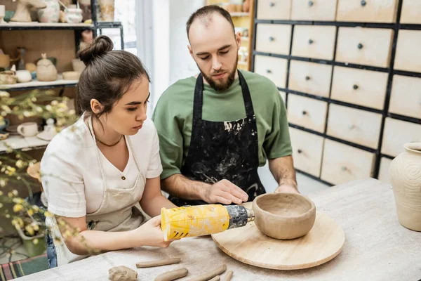 Couple of artisans in aprons drying clay bowl with heat gun while working in pottery workshop — Stock Photo