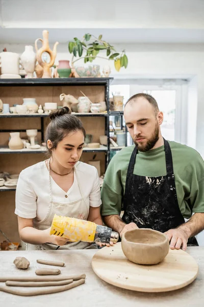 Couple of artisans in aprons drying clay bowl with heat gun on wooden board in ceramic workshop — Stock Photo