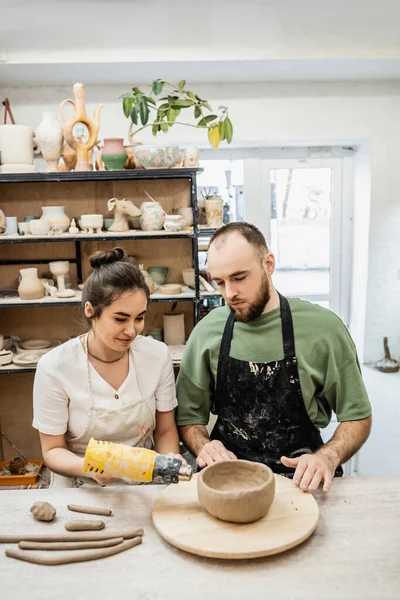 Smiling female artisan in apron drying ceramic bowl with heat gun near boyfriend in ceramic workshop — Stock Photo