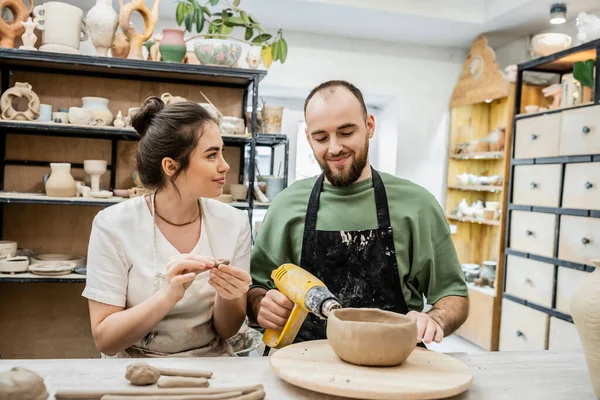Smiling craftswoman shaping clay while boyfriend drying bowl with heat gun in ceramic workshop — Stock Photo
