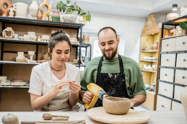 Casal positivo de artesãos em aventais trabalhando com argila e pistola de calor juntos na oficina de cerâmica — Fotografia de Stock