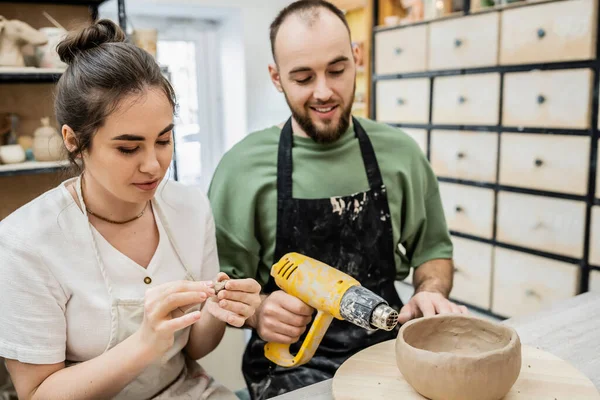 Lächelnde Kunsthandwerkerin formt Ton, während ihr Freund im Keramik-Atelier mit Heißluftpistole spricht — Stockfoto