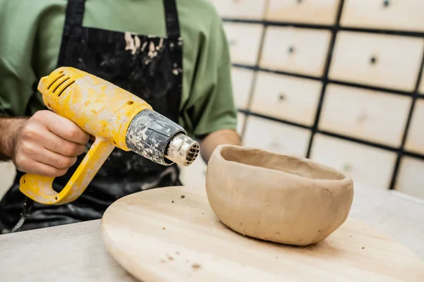 Обрезанный вид скульптора в фартуке сушки глиняной чаши с тепловым пистолетом на деревянной доске в керамической мастерской — стоковое фото