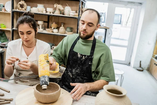 Couple d'artisans en tabliers travaillant ensemble avec pistolet à chaleur et de l'argile en atelier de poterie en arrière-plan — Photo de stock