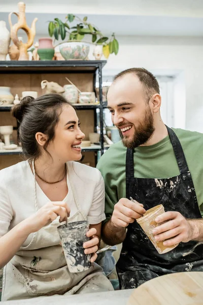 Smiling couple of artisans in aprons holding dye and paintbrushes while working in ceramic studio — Stock Photo
