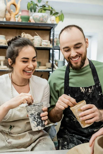 Couple souriant d'artisans tenant teinture et pinceaux près d'un bol d'argile sur un atelier de céramique — Photo de stock