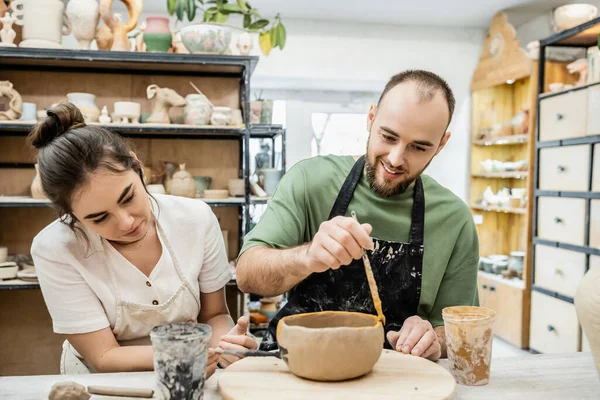 Smiling craftsman coloring clay bowl near girlfriend in ceramic workshop at background — Stock Photo