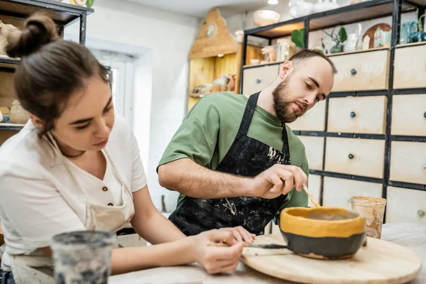 Bearded craftsman in apron coloring ceramic bowl with blurred girlfriend in ceramic workshop — Stock Photo