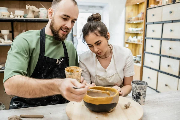 Smiling artisan in apron coloring clay bowl with girlfiend in ceramic workshop at background — Stock Photo