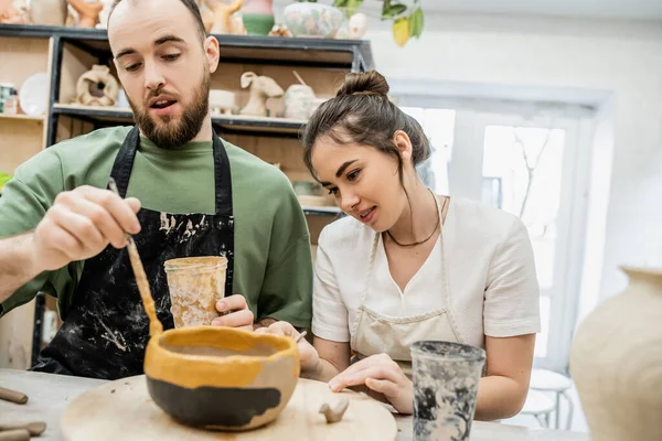 Artesana sonriente hablando con su novio mientras colorea tazón de barro en taller de cerámica en el fondo - foto de stock