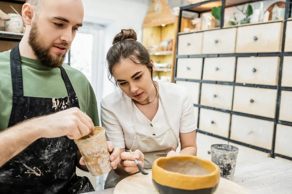 Ремесленник в фартуке раскрашивает глиняную чашу с размытой подругой вместе в керамической мастерской — стоковое фото