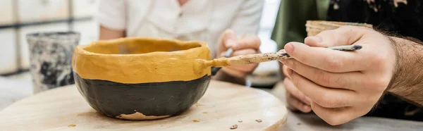 Smiling potters in aprons coloring ceramic bowl on wooden board in pottery studio at background — Stock Photo