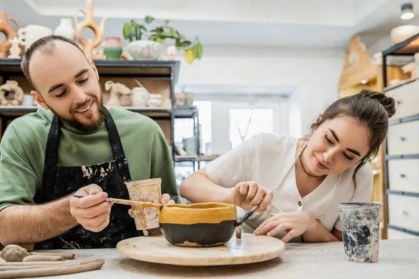 Cheerful craftspeople in aprons coloring clay bowl while working together in ceramic workshop — Stock Photo