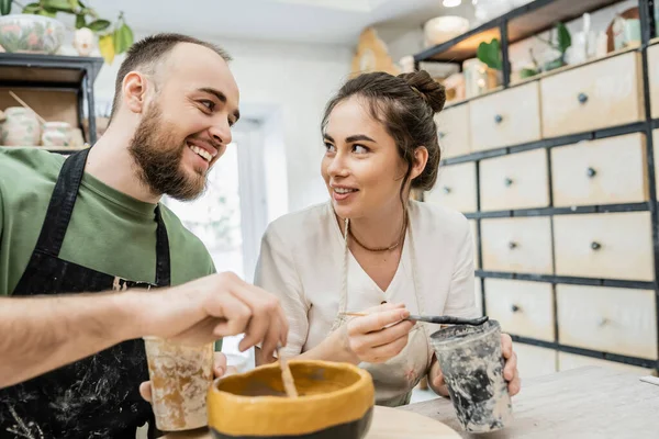 Artesãos românticos sorridentes em aventais falando e colorindo tigela de argila em oficina de cerâmica no fundo — Fotografia de Stock