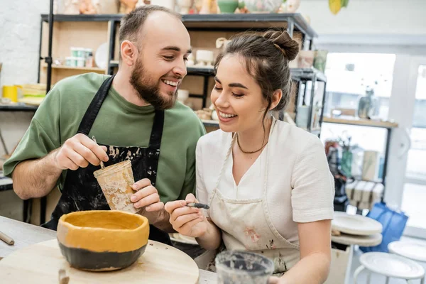 Cheerful couple of artisans holding paintbrushes whle coloring clay bowl on wooden board in studio — Stock Photo