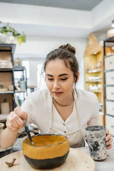 Brunette craftswoman holding plastic cup while dying clay bowl while working in pottery studio — Stock Photo
