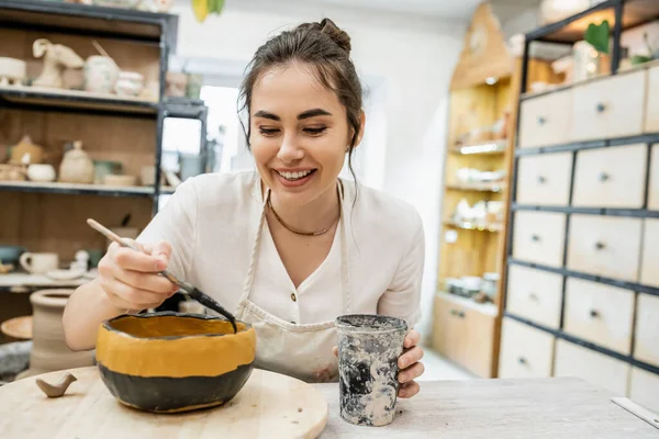 Glückliche brünette Handwerkerin Färbung Keramik Ton während der Arbeit in Töpferei Werkstatt im Hintergrund — Stockfoto