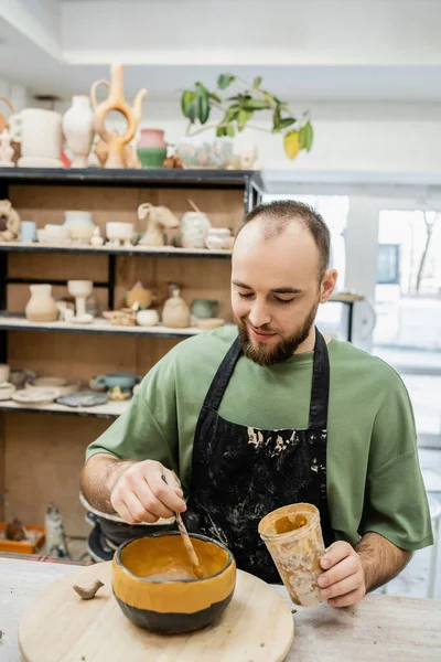 Радостный мужчина гончар в фартуке раскраски неостекленная глиняная чаша на деревянной доске в керамической студии — стоковое фото