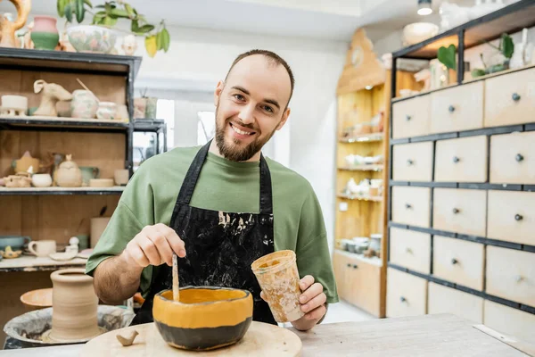 Smiling bearded craftsman in apron looking at camera and coloring clay bowl in pottery workshop — Stock Photo