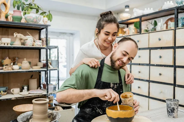 Artesã sorridente em avental abraçando namorado colorir tigela de argila em oficina de cerâmica no fundo — Fotografia de Stock